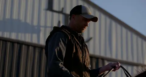 A farm worker in a cap holds a rope beside a corrugated metal barn.