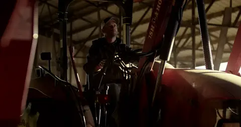 A farmer sits inside a red tractor cab in the dim light of a barn.