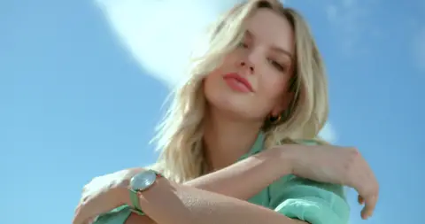 A blonde woman in a mint top poses against a bright blue sky in low-angle sunlight.