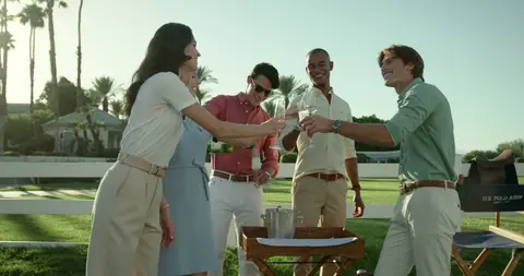 Four friends raise drinks around an outdoor table in bright afternoon light.