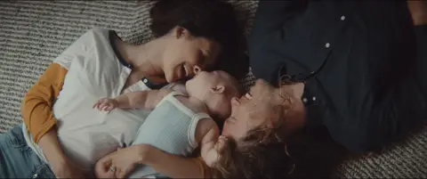 Two parents lie on a living room rug and cradle their baby between them.