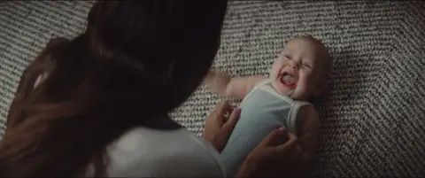 A laughing baby lies on a textured rug while a parent leans into the frame.