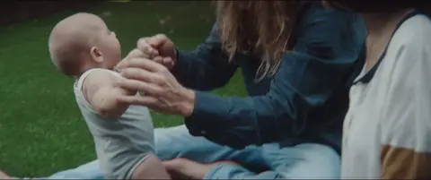 A baby reaches toward an adult's hands while being held outdoors on a lawn.