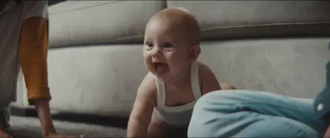 A smiling baby crawls on the floor beside a pale sofa in a softly lit home.