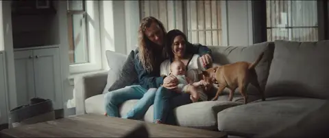 Two parents sit with their baby and dog on a sofa in a relaxed living room portrait.