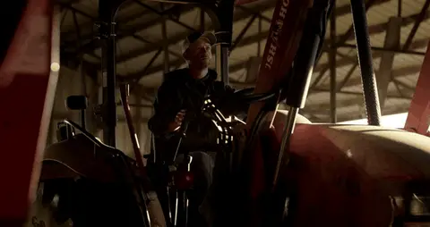 A farmer works beside a red tractor before moving toward an open barn doorway.