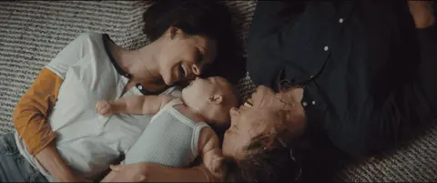 Two parents cuddle their baby on the floor before lifting the child together near a window.