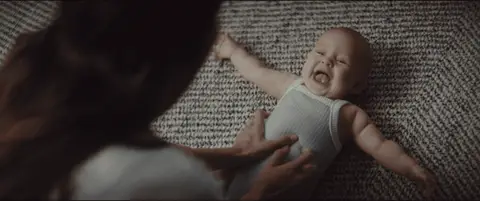 A baby laughs and rolls on a textured rug while a parent plays nearby.