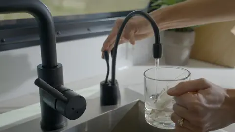 Strong interior lifestyle detail with faucet, water, glass, and hands showing use.