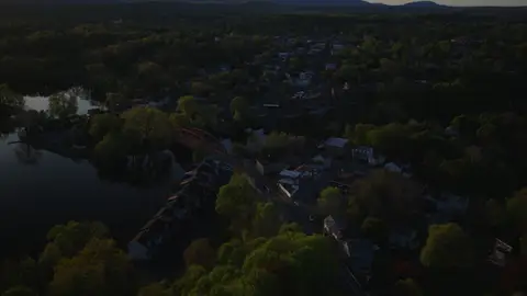 High aerial context shot of the river town, red bridge, lake or river bend, wooded shoreline, and distant hills.