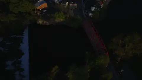Wide golden-hour aerial over a river village, red truss bridge, wooded shoreline, low sun, and mountain ridge.