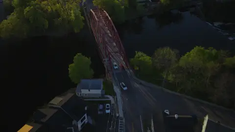 Top-down aerial of the red bridge, road, cars, nearby intersection, trees, and dark water.