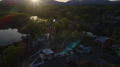 Wide golden-hour aerial over a river village, red truss bridge, wooded shoreline, low sun, and mountain ridge.
