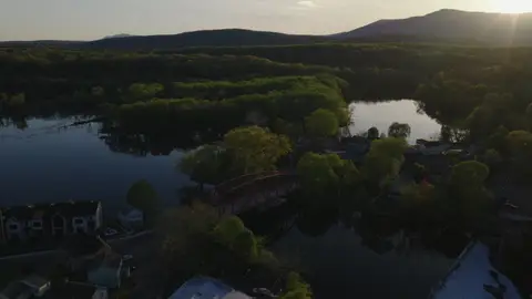 High, wide aerial over a river or lake valley, forest canopy, rural settlement, and mountain horizon at sunset.