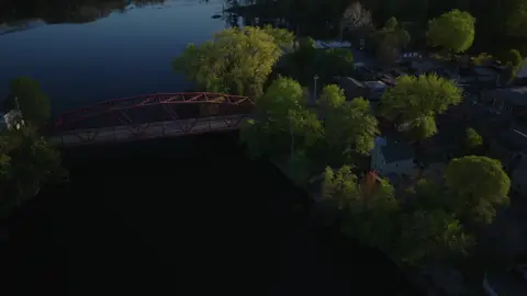 Aerial view of the red bridge beside a river dam or spillway, with wooded banks and small-town structures nearby.