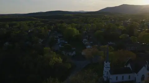 Backlit aerial over a white church steeple, cemetery, village rooftops, trees, and distant mountains.