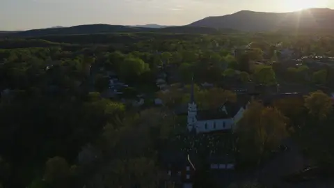 Backlit aerial over a white church steeple, cemetery, village rooftops, trees, and distant mountains.