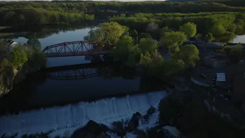Aerial view of the red bridge beside a river dam or spillway, with wooded banks and small-town structures nearby.
