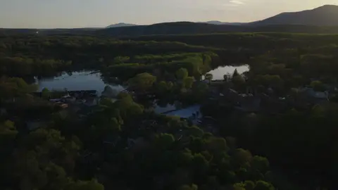 High, wide aerial over a river or lake valley, forest canopy, rural settlement, and mountain horizon at sunset.
