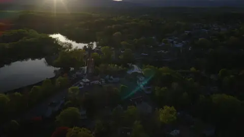 High aerial context shot of the river town, red bridge, lake or river bend, wooded shoreline, and distant hills.