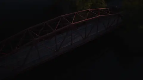 Wide golden-hour aerial over a river village, red truss bridge, wooded shoreline, low sun, and mountain ridge. Strong 6K source and scenic scale; heavy backlight, deep shadows, and lens flare need grading and crop review.