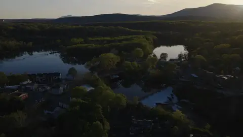 High, wide aerial over a river or lake valley, forest canopy, rural settlement, and mountain horizon at sunset. Good landscape context, but the shot is broad and subdued; may be too general unless the library needs wide regional establishing frames.