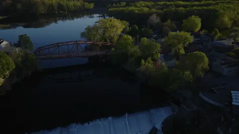 Aerial view of the red bridge beside a river dam or spillway, with wooded banks and small-town structures nearby. Clear subject combination with bridge and water feature; dusk contrast leaves some lower-frame areas dark.