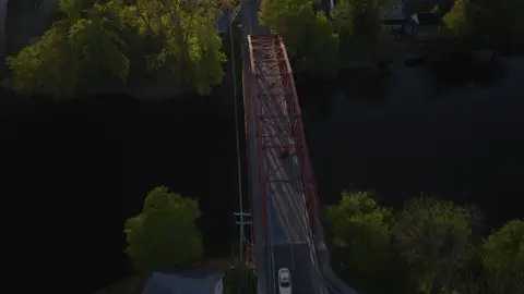 Top-down aerial of the red bridge, road, cars, nearby intersection, trees, and dark water. Graphic overhead composition, but the frame is shadow-heavy and vehicles are small; useful more as context than hero imagery.