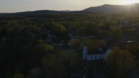 Backlit aerial over a white church steeple, cemetery, village rooftops, trees, and distant mountains. Attractive establishing frame, but the main subject is small and the low sun reduces shadow detail.