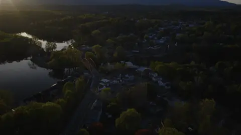 High aerial context shot of the river town, red bridge, lake or river bend, wooded shoreline, and distant hills. Strong context and scale; lower town details are dark, with flare and backlight requiring grade/crop review.