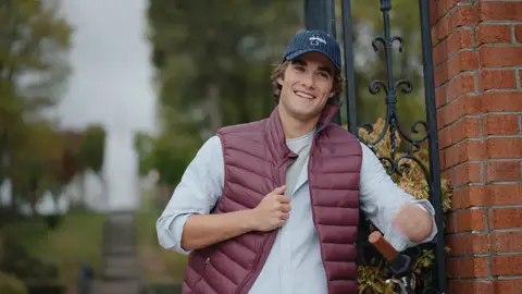 A man in a burgundy quilted vest and navy cap smiles beside a brick-and-iron garden gate.