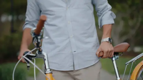 A bicycle handlebar and saddle sit beside a pale striped shirt, rolled cuff and resting hand at an iron gate.