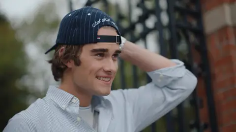 A man in a pale striped shirt smiles with one hand behind his backward navy cap beside an iron gate.