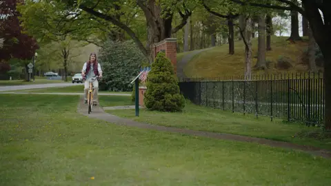A man in a burgundy vest rides a bicycle toward the camera along a curved path beside a black iron fence.