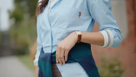 A wristwatch, rolled pale-blue shirt cuff and sweater tied at the waist are visible against a garden background.