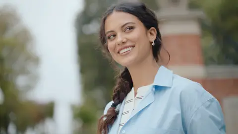 A woman with long braided hair smiles toward the camera in a pale blue overshirt and light striped top near the garden gate.