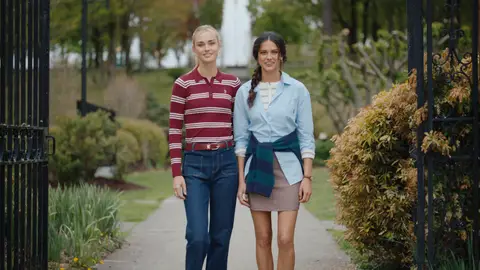 Two women stand together at an iron garden gate, one in a striped knit and the other in a pale blue shirt.