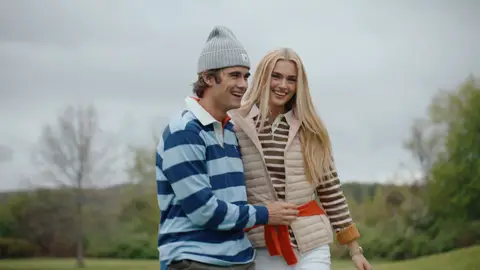 A man in a blue striped rugby shirt and gray beanie laughs with a woman in a cream quilted vest over a brown-and-cream striped top.