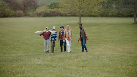 Five adults in layered casual wear move together across a wide green field.