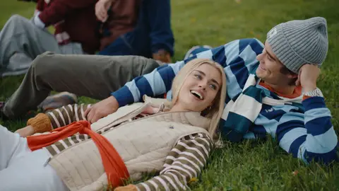 A reclining woman in a cream quilted vest looks upward laughing while a man in a blue striped rugby shirt and gray beanie smiles toward her.