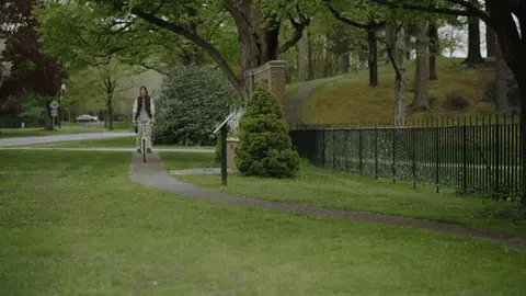 A man rides a bicycle toward the camera and follows the curve of a garden path beside an iron fence.