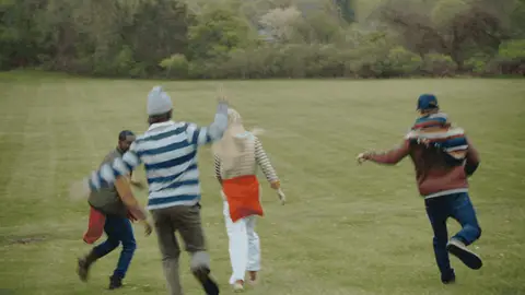 Four adults in layered outfits run away from the camera across a broad grassy field.