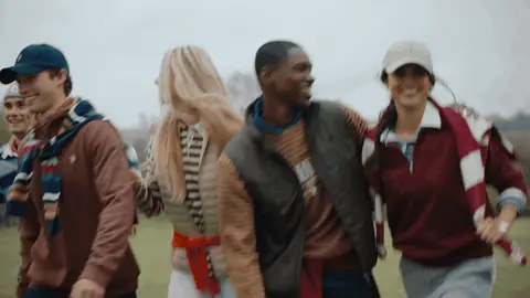 A close group of adults in layered apparel walks together and laughs across a grassy field.