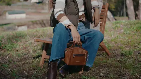 A seated person holds a brown leather handbag between blue-jean-clad knees, beneath a brown vest and striped sweater.