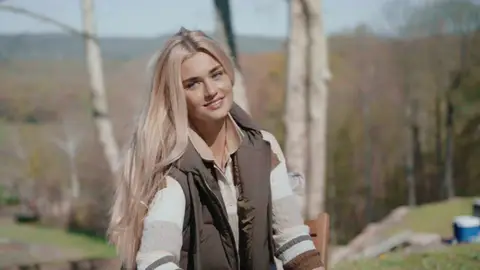 A blonde woman smiles while seated outdoors in a brown vest over a striped sweater, with trees and hills behind her.