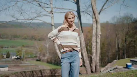 A blonde woman stands with a hand at her waist in a brown and cream striped sweater and blue jeans, with bare trees and hills behind her.