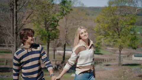 A woman in a cream and brown striped sweater holds hands with a man in a navy and tan striped shirt on a grassy hillside.