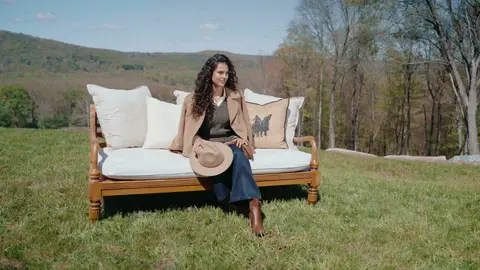 A woman in a camel blazer sits on a white-cushioned wooden sofa holding a brimmed hat in her lap, with grassy hills behind her.