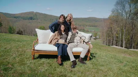 Two people sit on a white outdoor sofa while two others lean in behind them, with hills and trees in the background.