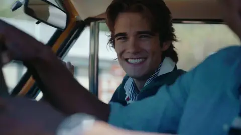 A smiling passenger with a striped collar is seen inside a vintage SUV, framed behind the driver and window opening.